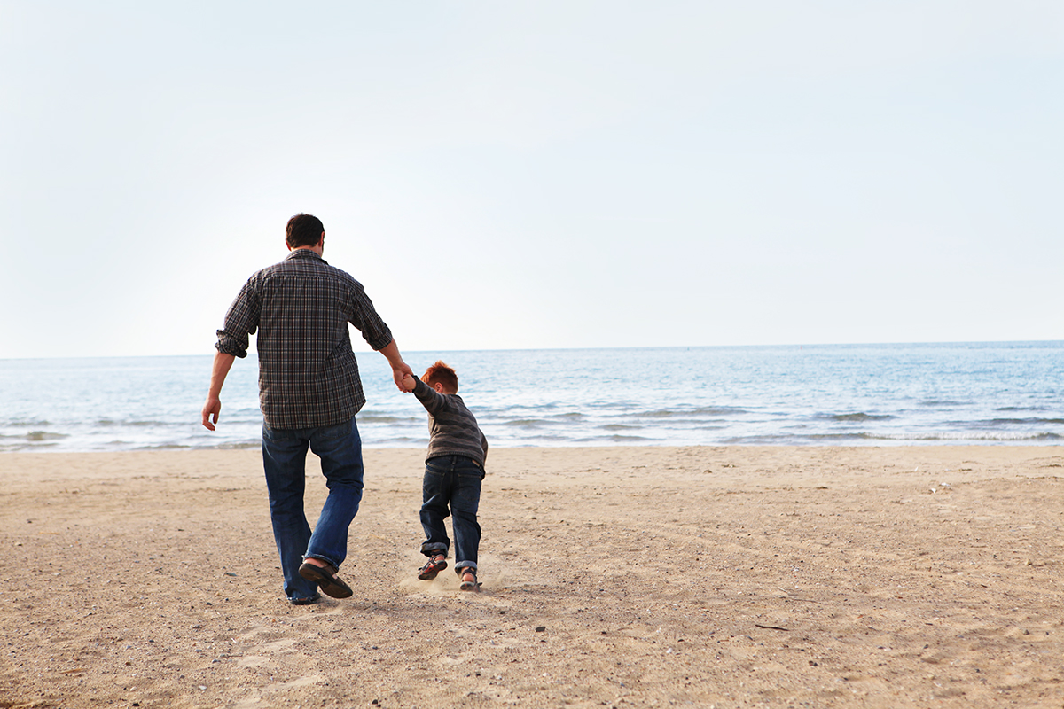 A dad holds his son's hand as they walk along the beach. Copy space.